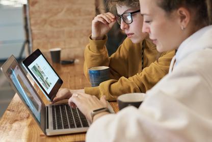 Two women in white and yellow sweatshirts talk and discuss the computer screens in front of them