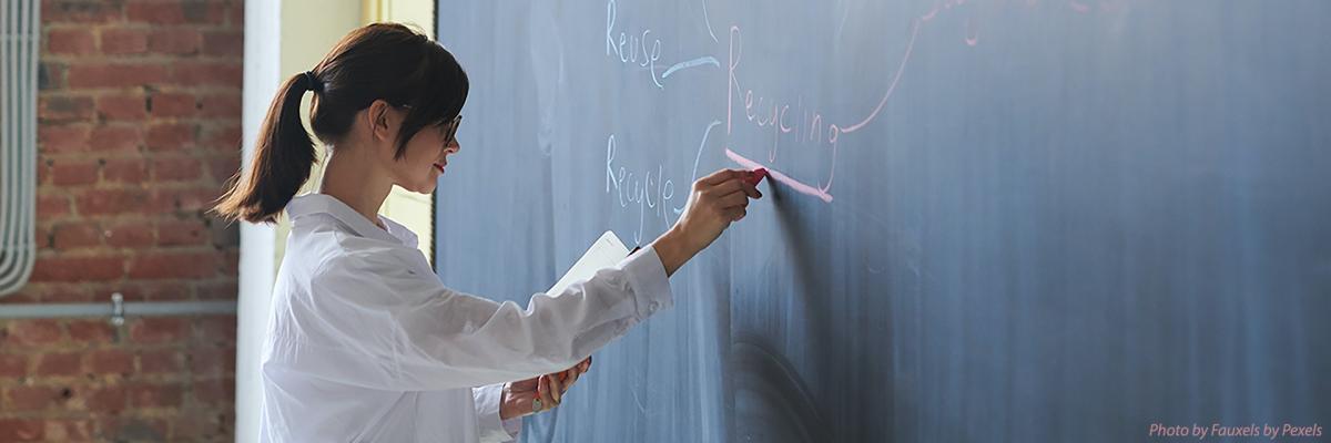 Femal instructor with her hair pulled back in a ponytail writes notes on a chalkboard as she teaches a class.